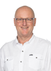 Lächelnder Mann mit Brille und kurzärmeligem weißen Hemd mit geknöpften Taschen, der vor einem schlichten Hintergrund steht.