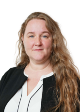 Eine Person mit langen, welligen Haaren lächelt leicht. Sie trägt eine schwarze Jacke über einem weißen Oberteil. Der Hintergrund ist schlicht.