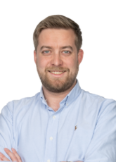 Ein Mann mit kurzen Haaren und Bart lächelt und trägt ein hellblaues Hemd. Seine Arme sind verschränkt, auf einem Unterarm ist ein Tattoo zu sehen. Der Hintergrund ist schlicht weiß.