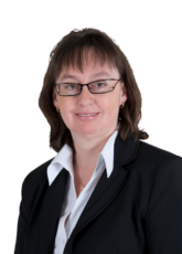 Eine Person mit schulterlangem braunem Haar und Brille trägt einen schwarzen Blazer und ein weißes Hemd mit Kragen und lächelt vor einem neutralen Hintergrund.