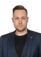 Eine Person mit kurzen Haaren und Bart, die einen dunklen Blazer über einem schwarzen Hemd trägt, blickt mit neutralem Gesichtsausdruck vor einem schlichten Hintergrund nach vorne.