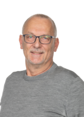 Ein älterer Mann mit kurzen grauen Haaren und Brille sowie einem grauen Hemd lächelt in die Kamera. Der Hintergrund ist schlicht weiß.
