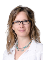 Frau mit welligem blondem Haar und Brille, trägt eine weiße Bluse und eine türkisfarbene Perlenkette und blickt vor einem weißen Hintergrund in die Kamera.
