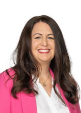 Eine Person mit langen, dunklen Haaren lächelt. Sie trägt einen leuchtend rosa Blazer über einer weißen Bluse. Der Hintergrund ist schlicht und hebt das Motiv hervor.