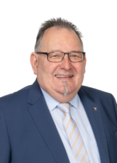 Ein Mann mit Brille, kurzen dunklen Haaren und Spitzbart lächelt. Er trägt einen blauen Anzug, ein hellblaues Hemd und eine gestreifte Krawatte. Der Hintergrund ist schlicht weiß.
