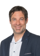 Ein Mann mit kurzen dunklen Haaren lächelt in die Kamera. Er trägt einen dunklen Blazer über einem weißen, gemusterten Hemd. Der Hintergrund ist schlicht weiß.