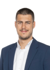 Ein Mann in blauem Blazer und weißem Hemd lächelt in die Kamera. Er hat kurze Haare und einen gestutzten Bart. Der Hintergrund ist schlicht weiß.