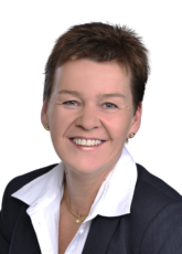 Lächelnde Person mit kurzen braunen Haaren, weißem Hemd mit Kragen und dunklem Blazer. Sie trägt goldene Ohrringe und eine Halskette vor einem schlichten Hintergrund.