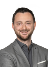 Eine Person mit kurzen dunklen Haaren und Bart lächelt in die Kamera. Sie trägt einen hellgrauen Blazer über einem dunklen Hemd und ein weißes Einstecktuch. Die Arme sind verschränkt. Der Hintergrund ist schlicht weiß.