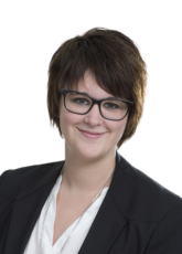 Eine Person mit kurzen, dunklen Haaren und Brille lächelt in die Kamera. Sie trägt einen schwarzen Blazer über einem weißen Hemd und eine silberne Halskette. Der Hintergrund ist neutral.