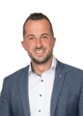 Mann mit kurzen Haaren und Bart, der ein weißes Hemd und einen blauen Blazer trägt, lächelt vor einem schlichten weißen Hintergrund.