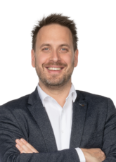 Ein Mann mit kurzen dunklen Haaren und Bart lächelt. Er trägt ein dunkles Jackett über einem weißen Hemd und hat die Arme verschränkt. Der Hintergrund ist schlicht und hell.
