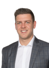 Ein Mann mit kurzen Haaren lächelt in die Kamera. Er trägt ein weißes Hemd und einen graukarierten Blazer. Der Hintergrund ist schlicht weiß.