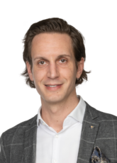 Eine Person mit mittellangem Haar trägt einen graukarierten Blazer, ein weißes Hemd und ein gemustertes Einstecktuch. Der Hintergrund ist schlicht weiß.