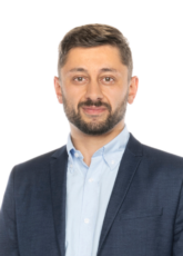 Ein Mann mit kurzen dunklen Haaren und Bart trägt ein hellblaues Hemd und ein dunkles Jackett. Er blickt nach vorne und lächelt leicht vor einem schlichten weißen Hintergrund.
