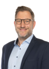 Ein Mann mit kurzen braunen Haaren, Bart und Brille lächelt. Er trägt einen marineblauen Blazer über einem hellblauen Hemd vor einem schlichten weißen Hintergrund.