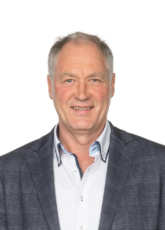 Mann mit kurzen grauen Haaren, trägt ein blaues Jackett und ein weißes Hemd und lächelt vor einem schlichten Hintergrund.