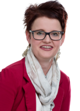 Eine Person mit kurzen dunklen Haaren und Brille lächelt in die Kamera. Sie trägt einen roten Blazer und einen hellen Schal vor einem schlichten Hintergrund.