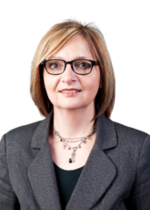 Eine Frau mit schulterlangem Haar und Brille trägt einen dunklen Nadelstreifenblazer und eine Kette mit kleinen Perlen. Sie lächelt leicht vor einem schlichten Hintergrund.