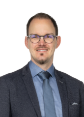 Ein Mann mit Brille, blauem Hemd, grauer Krawatte und dunklem Blazer mit Strukturmuster lächelt vor einem weißen Hintergrund in die Kamera.
