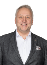 Lächelnder Mann mit kurzen grauen Haaren, der ein dunkles Jackett, ein weißes Hemd und ein gemustertes Einstecktuch trägt und vor einem schlichten Hintergrund steht.
