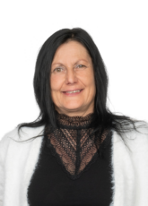 Lächelnde Frau mit langen schwarzen Haaren, die vor einem schlichten Hintergrund ein schwarzes Spitzentop und eine weiße Strickjacke trägt.