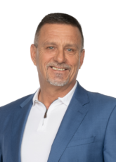 Lächelnder Mann, der einen hellblauen Blazer über einem weißen Hemd trägt. Er hat kurze Haare und einen Spitzbart. Der Hintergrund ist weiß.