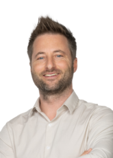 Ein lächelnder Mann mit kurzen, braunen Haaren und Bart steht mit verschränkten Armen da. Er trägt ein helles Hemd. Der Hintergrund ist schlicht weiß.