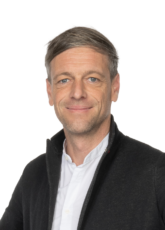 Ein Mann mit kurzen grauen Haaren und hellem Bartstoppeln lächelt leicht. Er trägt ein weißes Hemd und eine dunkle Jacke. Der Hintergrund ist schlicht weiß.