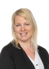 Eine Frau mit schulterlangem blondem Haar lächelt in die Kamera. Sie trägt einen schwarzen Blazer über einer weißen Bluse und kleine Ohrringe. Der Hintergrund ist schlicht weiß.