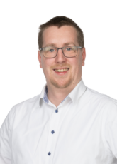 Lächelnde Person mit Brille und weißem Hemd mit blauen Knöpfen, die vor einem schlichten Hintergrund steht.