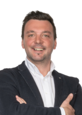 Lächelnder Mann mit kurzen dunklen Haaren, weißem Hemd und dunklem Blazer, steht mit verschränkten Armen vor einem weißen Hintergrund.