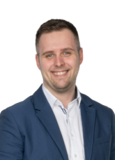 Ein Mann mit kurzen braunen Haaren, der einen blauen Blazer über einem weißen Hemd trägt, lächelt. Der Hintergrund ist schlicht und hell, was seinen freundlichen Ausdruck unterstreicht.