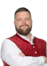 Ein bärtiger Mann mit kurzen Haaren, der ein weißes Hemd und eine rote Weste mit einem komplizierten Muster trägt, verschränkt die Arme und lächelt selbstbewusst.