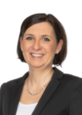 Eine Frau mit kurzen braunen Haaren lächelt in die Kamera. Sie trägt einen schwarzen Blazer über einem weißen Top und eine Perlenkette. Der Hintergrund ist schlicht weiß.