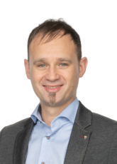 Lächelnder Mann mit kurzen dunklen Haaren, hellblauem Hemd und dunklem Blazer. Er hat einen kleinen Spitzbart und steht vor einem schlichten weißen Hintergrund.