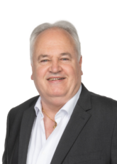 Ein lächelnder Mann mit kurzen weißen Haaren trägt ein dunkles Jackett über einem weißen Hemd und eine goldene Halskette. Der Hintergrund ist schlicht weiß.