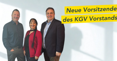 Drei Personen stehen lächelnd da, zwei Männer und eine Frau. Sie tragen halbformelle Kleidung. In der oberen rechten Ecke befindet sich ein gelbes Banner mit der Aufschrift „Neue Vorsitzende des KGV-Vorstands“.