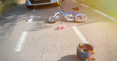 Ein Kinderfahrrad liegt auf einer asphaltierten Straße. Helm und Schuhe liegen verstreut herum. Im Hintergrund parkt ein weißes Auto, das auf eine mögliche Unfallstelle hindeutet. Das Bild ist sonnig und wirft weiche Schatten auf den Boden.